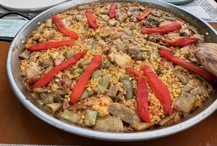 Una paella grande y plana llena de arroz amarillo con trozos de pollo, judía ancha verde y tiras de pimiento rojo por encima; a la derecha, una cuchara de madera y, al fondo, platos en la mesa.