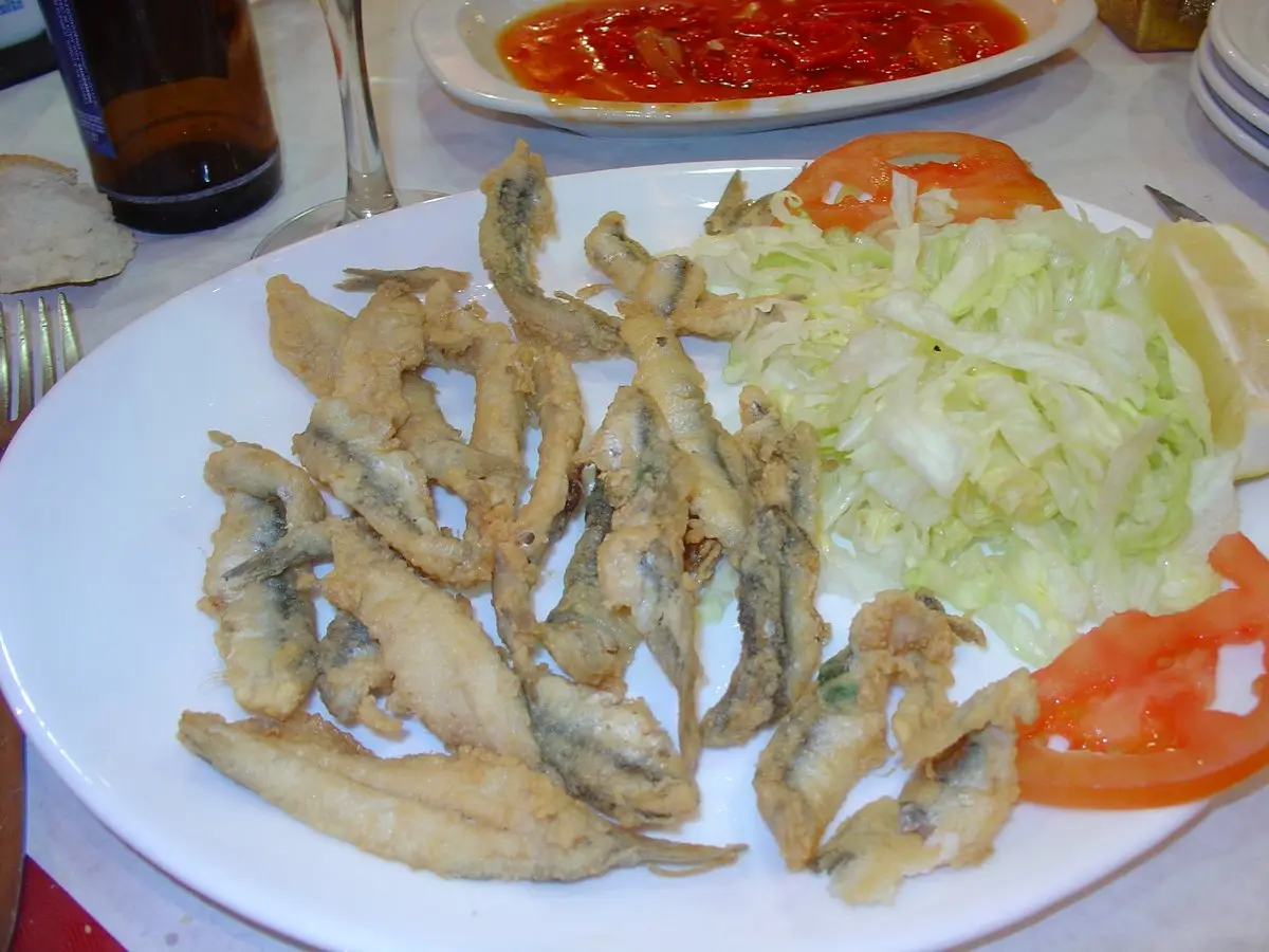 Un plato blanco con una docena de pescaditos fritos rebozados, junto a lechuga en tiras, rodajas de tomate y un gajo de limón; al fondo, una fuente con salsa roja y una copa.