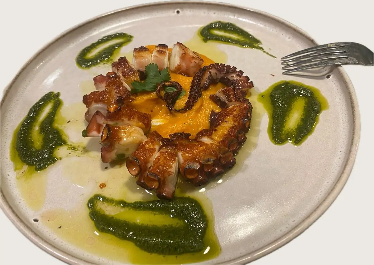 Un plato claro de gres con patas de pulpo a la plancha dispuestas en círculo sobre un puré anaranjado, con cilantro; alrededor, toques de salsa verde de hierbas y aceite de oliva, y un tenedor a la derecha.