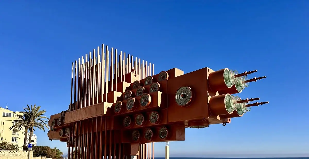 Una escultura de acero rojizo contra un cielo sin nubes: un bloque horizontal del que sobresalen cilindros con discos de vidrio, encima una cresta de barras verticales y a la derecha el mar