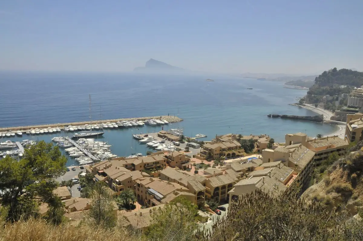Vista desde la ladera sobre un puerto deportivo: pantalanes llenos de barcos tras un dique largo y, delante, la urbanización ocre; al fondo, el peñón de Calp entre la calima