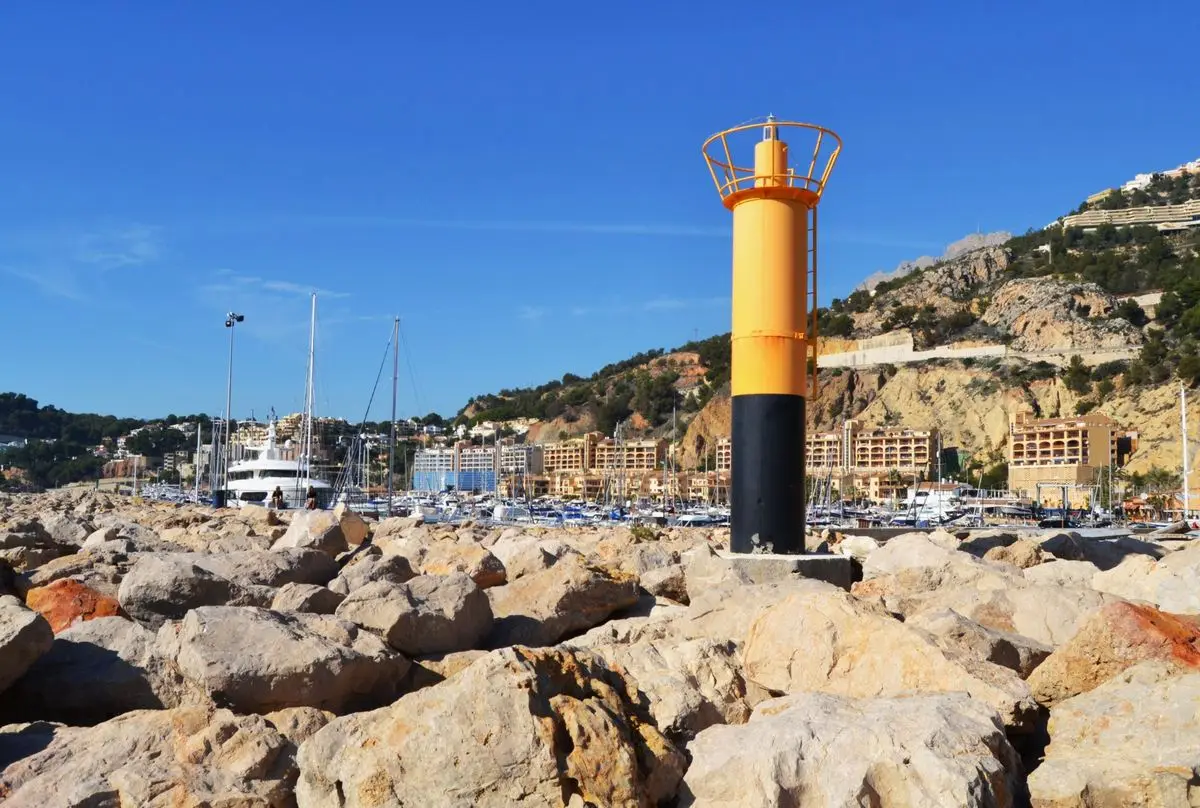 Grandes bloques de escollera clara en primer término, con un faro naranja y negro encima; detrás, el bosque de mástiles del puerto, la urbanización ocre y el talud cortado