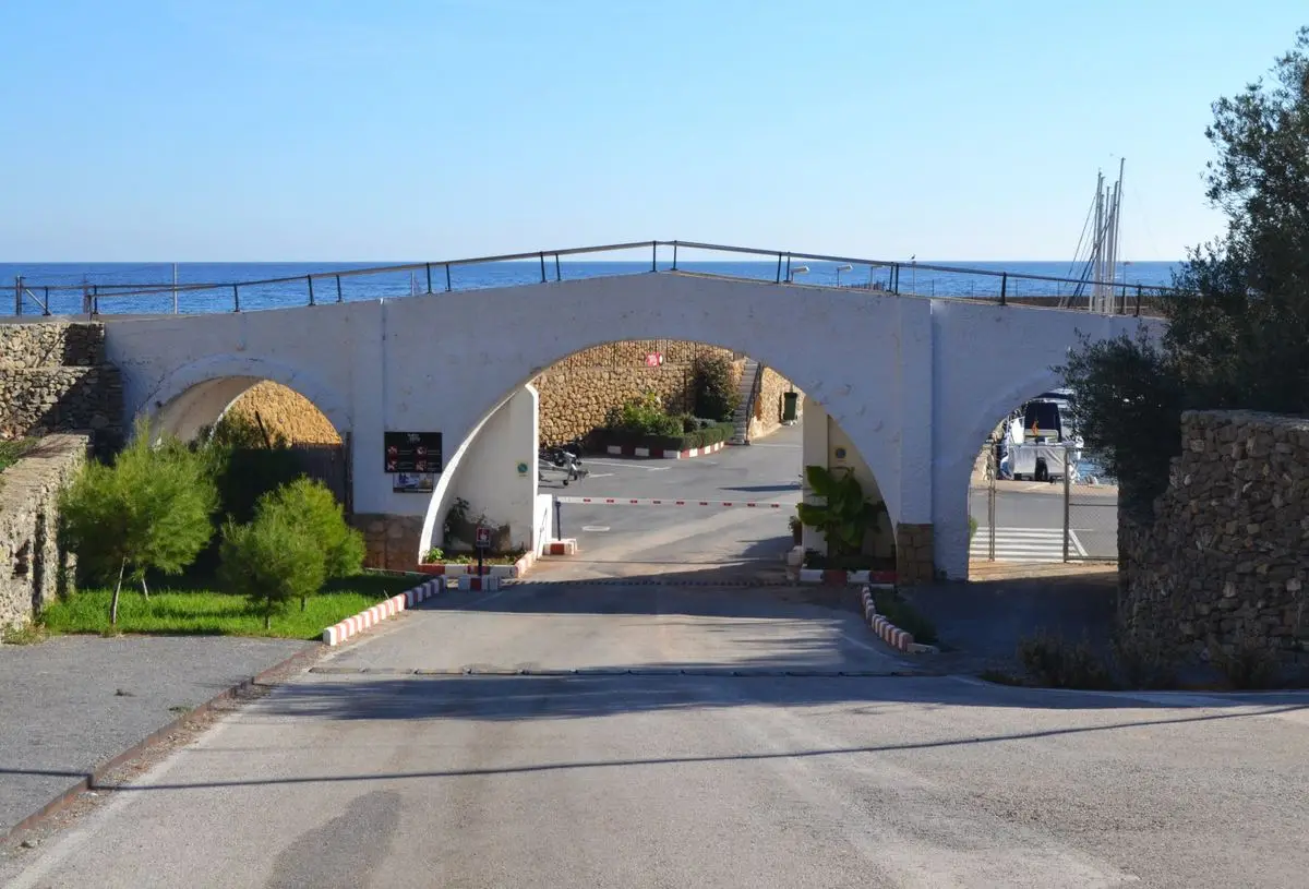 Un puente de arcos encalado sobre el acceso al puerto, con tres vanos, una barandilla arriba y el mar abierto detrás; debajo, la barrera, el aparcamiento y los mástiles