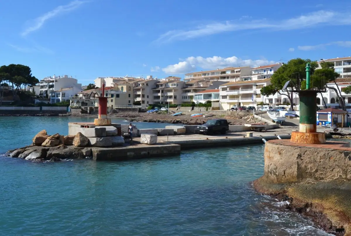 La bocana de un puerto pequeño entre dos espigones cortos, con un faro en cada uno —rojo a la izquierda, verde a la derecha—; detrás, la rampa, las viviendas y la ladera arbolada