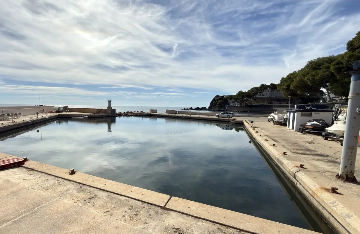 Pequeña dársena rectangular de agua en calma que refleja nubes finas, entre muelles de hormigón. A la izquierda, una terraza con sombrillas; al fondo, el espigón y el mar; a la derecha, motos de agua, una caseta de aseos y pinos.