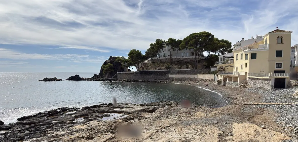 Pequeña cala con losas de roca planas y arena en primer plano, agua tranquila y una playa de guijarros que lleva a la derecha hasta una casa amarilla con torrecilla. A la izquierda, un promontorio con pinos y casas; contraluz sobre el mar.
