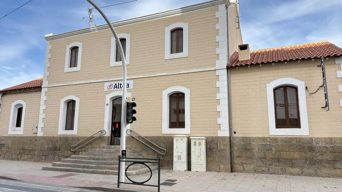 Estación de dos plantas color arena, con ventanas en arco enmarcadas en blanco y zócalo de piedra; sobre la puerta, el letrero «Altea» con un logotipo naranja «T». Delante, un semáforo, una escalera con barandillas y dos armarios eléctricos.