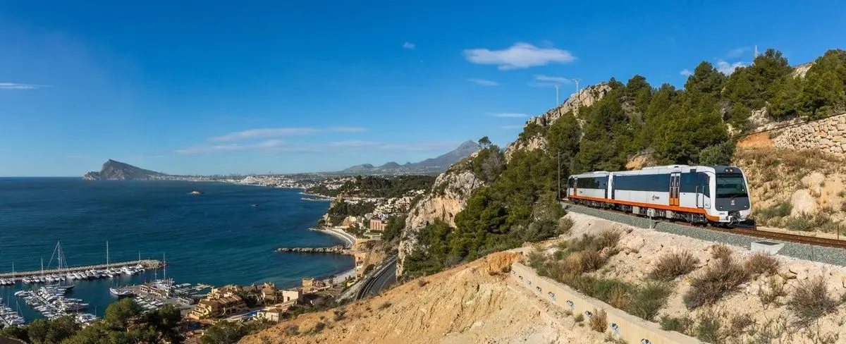Una unidad blanca y naranja por la vía costera, muy por encima de un puerto deportivo; al fondo, la bahía y Benidorm