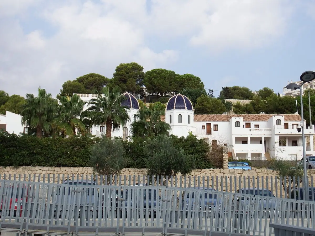 Dos cúpulas de escama azul vidriada con nervios claros sobre una casa blanca de ventanas de medio punto, con palmeras y pinos delante; en primer término, un muro de piedra seca y la valla de un aparcamiento