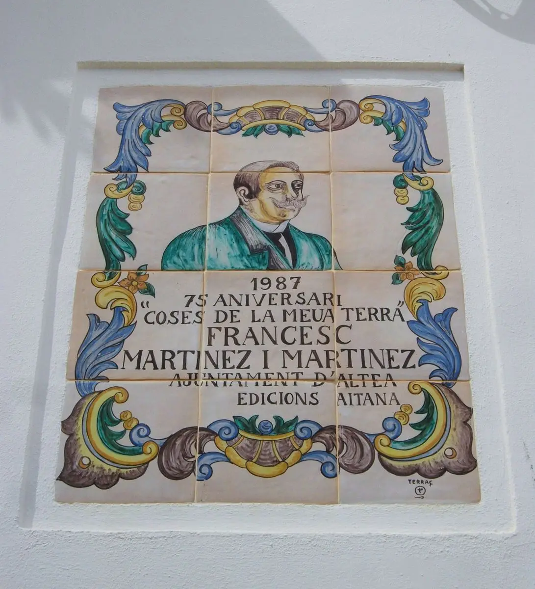 Un panel cerámico de doce azulejos pintados a mano con orla floral en azul, verde y amarillo; en el centro, el busto de un hombre con bigote y, debajo, la inscripción 1987, 75 aniversari, Coses de la meua terra, Francesc Martínez i Martínez