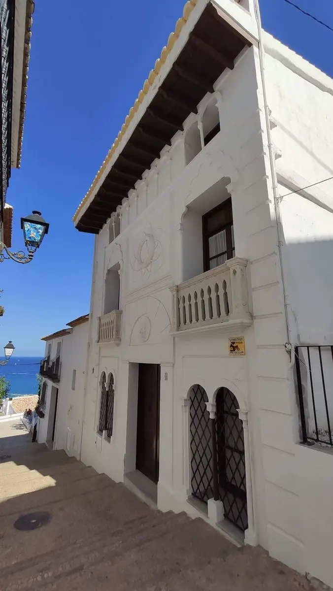 La fachada blanca de la Casa Cervantes en una calle en pendiente: arcos de herradura, rosetones de yeso y dos balcones de balaustres torneados; abajo a la izquierda, el mar
