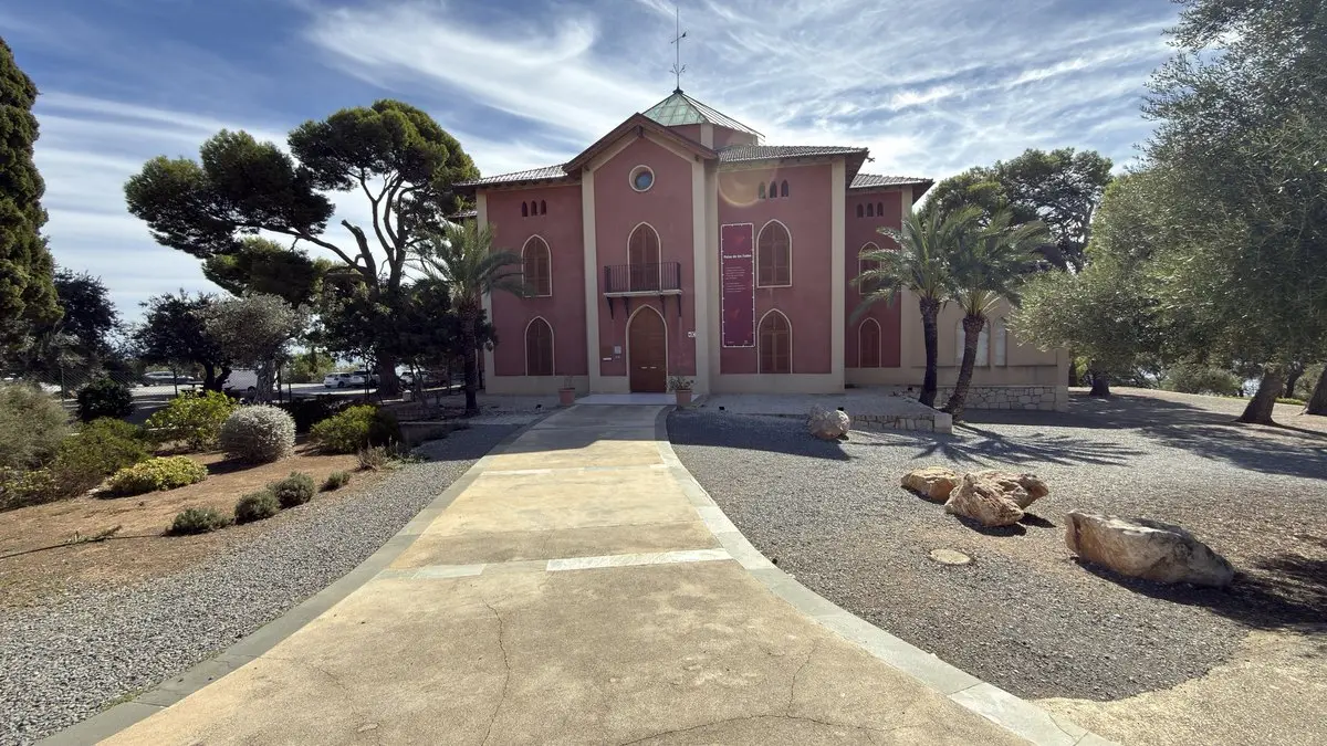 Villa de dos plantas en rosa rojizo, de estilo historicista, con ventanas apuntadas, pequeño balcón y linterna verde en el tejado; en la fachada, una pancarta «Palau de les Fades». Un ancho camino cruza zonas de grava con rocas; pinos, palmeras y olivos alrededor.