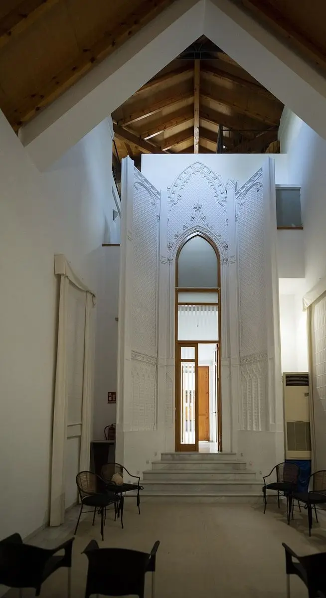 Un interior alto y blanco con armadura de madera vista; en el centro, una portada apuntada con fino trabajo de yeso neogótico, con peldaños y sillas delante
