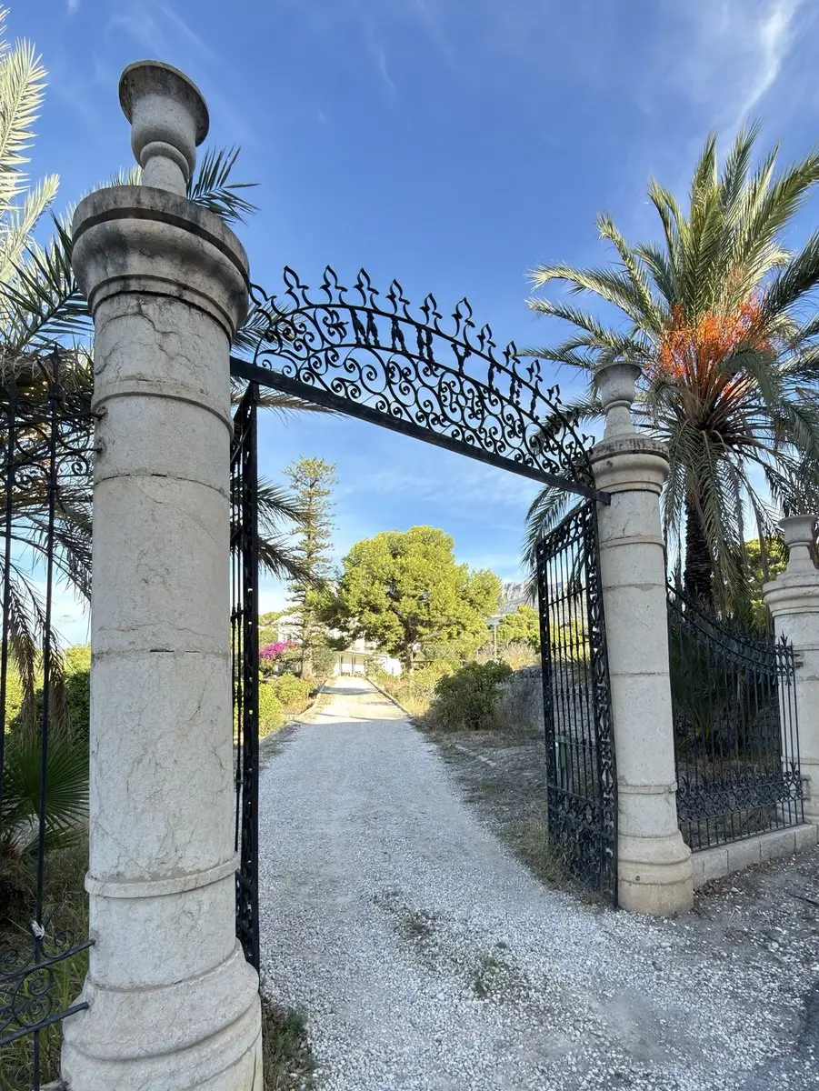 Portón de hierro forjado entre dos columnas de piedra clara rematadas con jarrones; en el arco, la inscripción «MARYCEL». Un camino de grava lleva entre palmeras y arbustos hasta una casa blanca al fondo, bajo cielo azul.