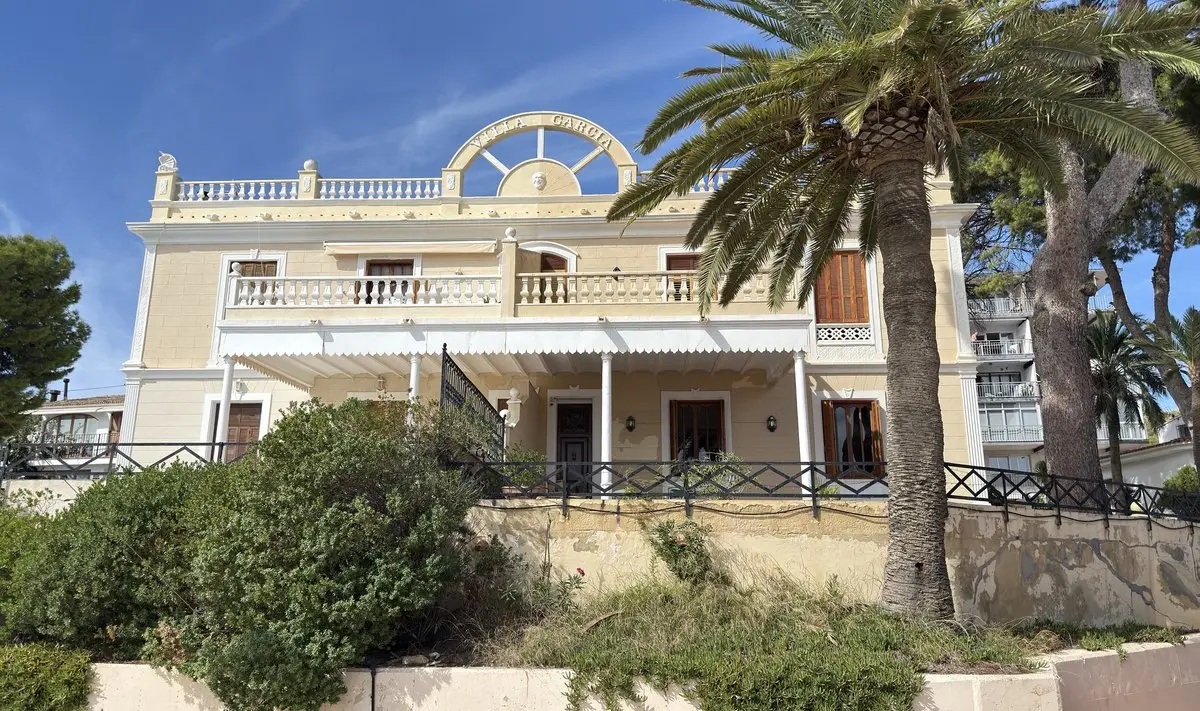 Villa color crema de dos plantas con balaustradas, porche y contraventanas de madera; en la cubierta, un remate en arco con la inscripción «VILLA GARCIA» y un relieve de rostro. A la derecha, una alta palmera datilera; delante, jardín frondoso y un muro desconchado.